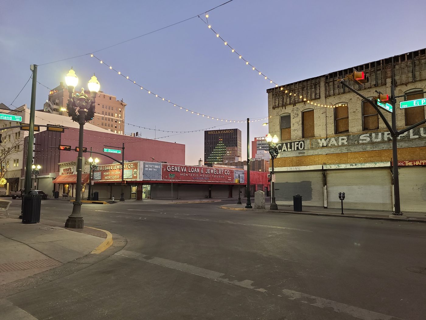 An empty downtown El Paso street corner at dawn, strung with lights, with an old war surplus store on the corner